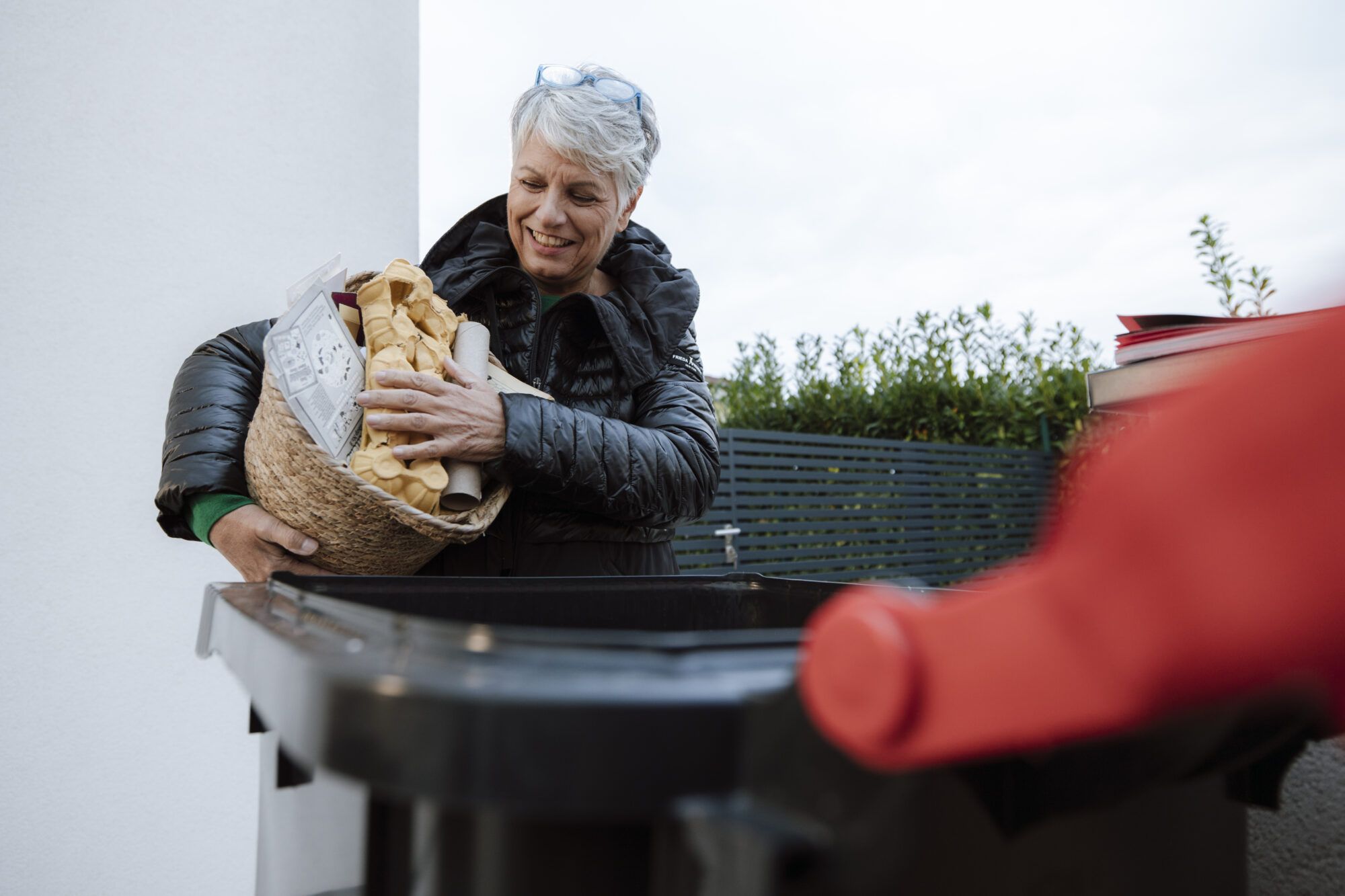 Eine Frau mit schwarzem Mantel und grauen Haaren hält einen Korb gefüllt mit Altpapier wie Eierkartons oder Zeitschriften in der Hand. Vor der Frau ist eine schwarze Tonne mit rotem Deckel geöffnet zu erkennen.