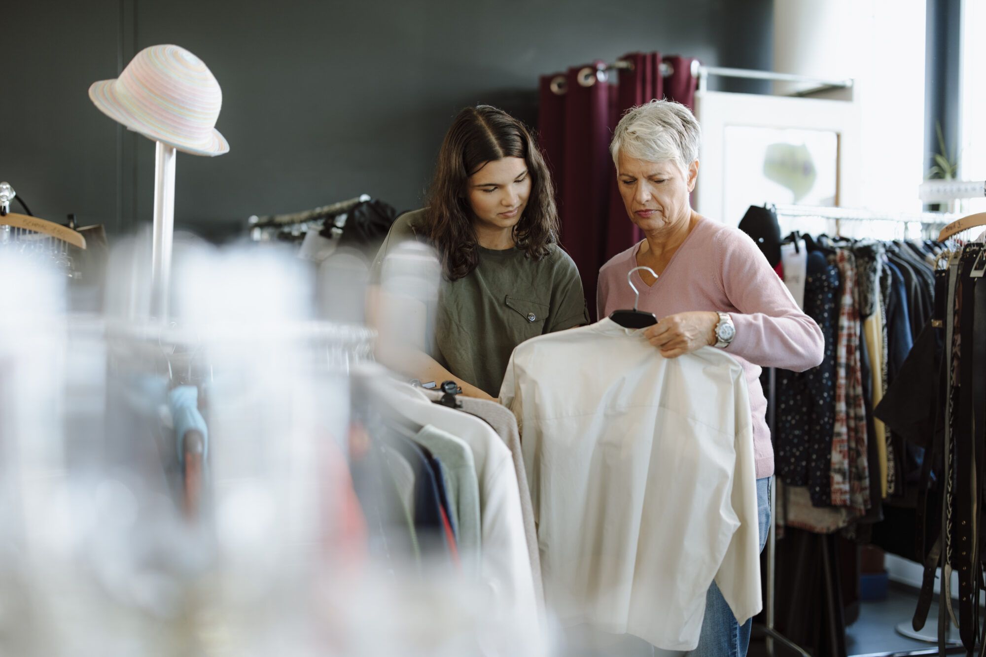 Eine ältere Dame mit grauem Haar steht in einem Secondhandladen und hält eine weiße Bluse in der Hand. Neben ihr steht ein junges Mädchen mit lockigem Haar und einem olivgrünen T-Shirt, das ebenfalls die Bluse interessiert betrachtet.