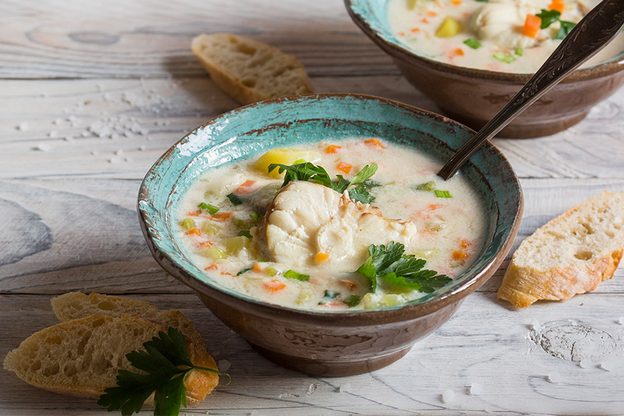 Braun-türkise Keramikschüssel gefüllt mit cremigem Fischeintopf. Zu sehen sind Karottenstücke, Fischstücke und Kräuter. Neben der Schüssel liegen Weißbrotschnitten auf einem grauen Holztisch.