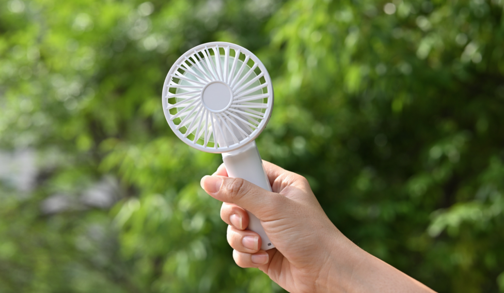 Zu sehen in eine Hand, die einen wei&szlig;en Handventilator h&auml;lt. Im Hintergrund ist Natur zu sehen.