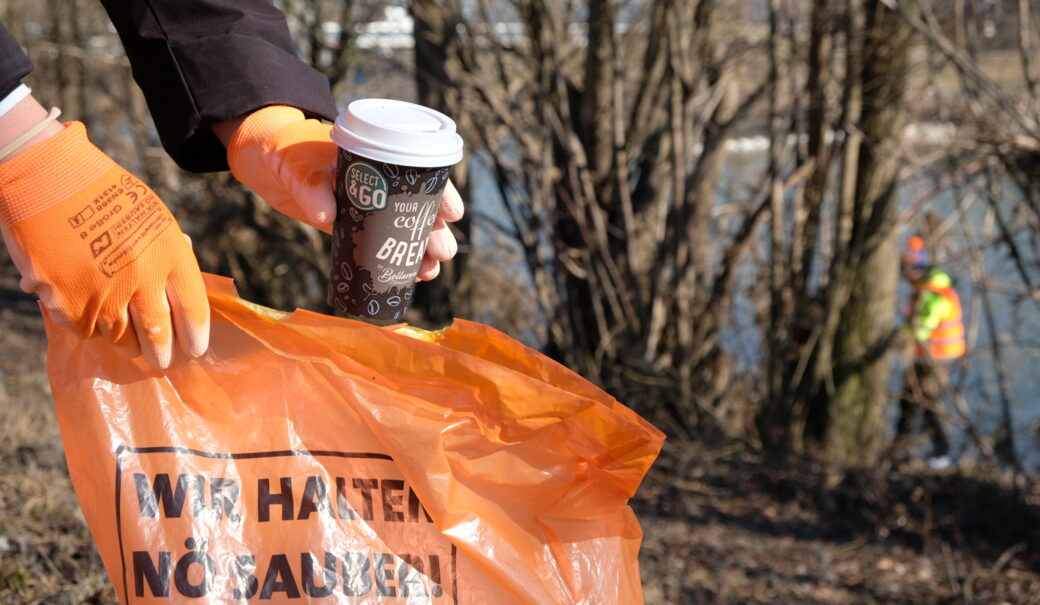 Auf dem Bild ist eine Person zu sehen, die mit orangen Handschuhen einen Coffee-to-go-Becher aufnimmt und in einen orangenen M&uuml;llsack wirft. Im Hintergrund sind weitere Personen in Warnwesten an einem Flussufer besch&auml;ftigt, offenbar bei einer Fluss- oder Uferreinigungsaktion. Auf dem M&uuml;llsack steht: &bdquo;WIR HALTEN N&Ouml; SAUBER!&ldquo;.
