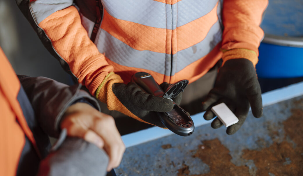Man sieht einen Ausschnitt von einem WSZ Mitarbeiter, der eine orange Jacke trägt und ein elektronisches Gerät von einem Akku trennt und beide jeweils in einer Hand hält.