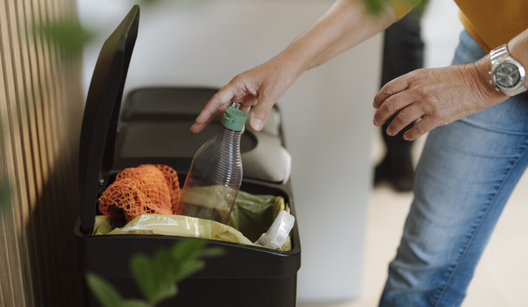 Auf dem Foto sieht man einen Müllstation für drei unterschiedliche Abfälle. Eine Frau entsorgt gerade eine leere Reinigungsflasche in den Behälter für Verpackungsabfälle.